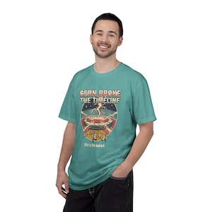May include: A teal short-sleeve t-shirt featuring a graphic print of a circular particle accelerator with a lightning bolt striking it. The text reads CERN BROKE THE TIMELINE and this is the bad one, with small figures running away from the structure.