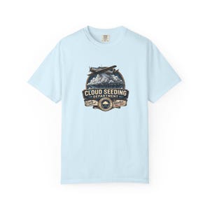 May include: A light blue short-sleeve t-shirt featuring a graphic of an airplane flying over clouds. The text reads CLOUD SEEDING DEPARTMENT, Locally Sourced, and Weather Since 1947. The design includes a circular emblem with a rain cloud icon.