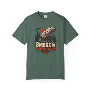May include: A sage green short-sleeve t-shirt featuring a graphic print of a server farm building emitting smoke into a forest landscape under a red sun and satellite. The text reads SMOKE & SERVER FARMS with a small fire warning triangle icon.