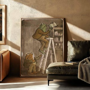 Peut inclure: Une impression d'art encadrée représentant une grenouille verte debout sur une échelle en bois pour atteindre une pile de livres sur une étagère haute, tandis qu'une autre grenouille verte se tient au sol, portant des vestes et pantalons marron.
