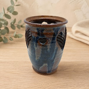 May include: A cylindrical ceramic vessel featuring a glossy glaze in shades of deep blue and earthy brown. The exterior surface is punctuated by several heart-shaped cutouts, revealing the interior space of the pottery piece.