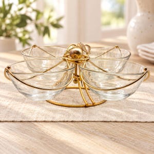 May include: A gold-toned metal serving stand featuring four clear glass bowls arranged in a circular pattern. The central structure is adorned with a decorative gold pear and leaf motif, supported by a circular base with curved metal arms.