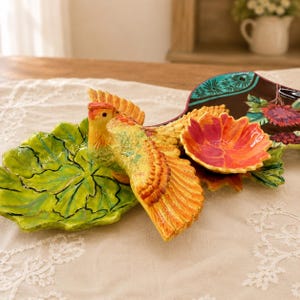 May include: A ceramic serving dish shaped like a bird with outstretched wings, featuring a bright yellow and orange body, a green leaf-shaped section, and a separate small bowl with a vibrant pink and orange flower petal design.