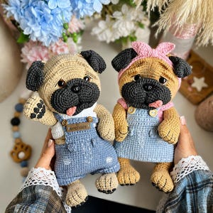 Peut inclure: Deux poupées carlin au crochet avec un corps beige et un visage noir. Le carlin de gauche porte une salopette en jean bleu avec une étiquette HAND MADE. Le carlin de droite porte un bandeau rose et une robe en jean bleu à boutons jaunes.