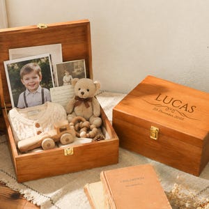 May include: Two rectangular wooden keepsake boxes with gold-tone metal latches. One box is open, containing a photograph of a child, a beige knit sweater, a small teddy bear, and a wooden toy truck. The closed box is engraved with the name LUCAS.