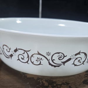 May include: A white oval casserole dish made of glass featuring a dark brown decorative pattern of swirling vines and small floral accents around the exterior. The dish includes integrated handles on both sides and a smooth, glossy finish.