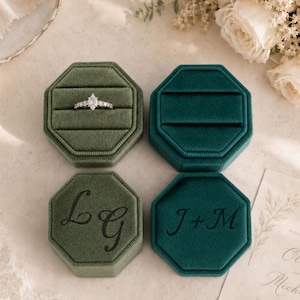 Op de afbeelding: Twee achthoekige fluwelen ringdoosjes in olijfgroen en diepblauwgroen. Het olijfgroene doosje bevat een ring met een marquise-geslepen diamant en het monogram LG. Het blauwgroene doosje is leeg en heeft het monogram J+M. Beide hebben bijpassende deksels.