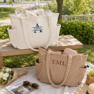 May include: Two corduroy tote bags with side pockets and embroidered personalisation. The cream bag features a blue monogram with the name Ashley, while the tan bag displays the brown embroidered initials EML. Both bags have dual handles.