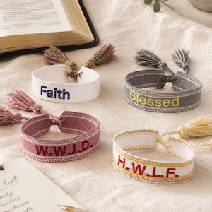 May include: Four woven fabric bracelets featuring embroidered text and decorative tassels. One white bracelet reads Faith, a gray one reads Blessed, a dusty rose one reads W.W.J.D., and a white one with yellow trim reads H.W.L.F. in red thread.