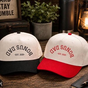 Op de afbeelding: Twee baseballpetten met gebroken witte kronen en gestructureerde kleppen. De ene pet heeft een zwarte klep en zwart geborduurde tekst BONUS DAD -EST. 2026-. De andere pet heeft een rode klep en rood geborduurde tekst BONUS DAD -EST. 2026-.