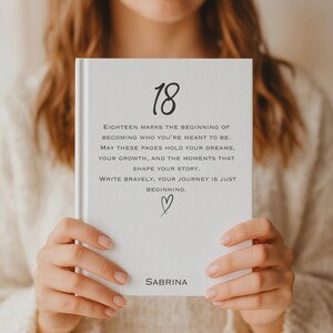 May include: A white hardcover journal featuring the number 18 in a large script font at the top. Below, the text reads: Eighteen marks the beginning of becoming who you're meant to be. May these pages hold your dreams, your growth, and the moments that shape your story. Write bravely, your journey is just beginning. A small heart icon is centered below the text, and the name Sabrina is printed at the bottom.