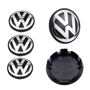 May include: A set of four circular wheel centre caps featuring the silver and black Volkswagen logo on the front. The reverse side of one cap is black, displaying moulded text including MADE IN GERMANY and various technical part markings.