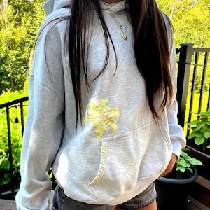 May include: A light gray hooded sweatshirt featuring a yellow embroidered lily flower design on the front, positioned above a kangaroo pocket with yellow decorative stitching along the pocket edge, paired with dark gray denim shorts.
