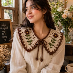 May include: A cream-colored long-sleeved blouse featuring a detachable crochet collar. The collar is beige with a dark brown scalloped edge and embroidered brown flowers with green stems. It includes a brown crochet tie with small floral accents.