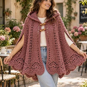 May include: A dusty rose crochet hooded cape featuring an intricate openwork pattern, a scalloped hem with circular motifs, and two light brown wooden toggle closures at the neckline. The garment is worn over a white top and light blue denim jeans.