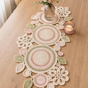 May include: A long, decorative crochet table runner featuring a series of interconnected circular mats, flower-shaped motifs, and leaf-shaped accents in soft shades of cream, sage green, and dusty pink, arranged in a continuous, organic pattern.