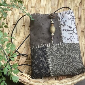 May include: A rectangular fabric pouch featuring a patchwork design of brown, grey, and white patterned textiles joined by visible white stitching. A decorative charm with wooden and metallic beads hangs from the top, attached to a thin black cord.