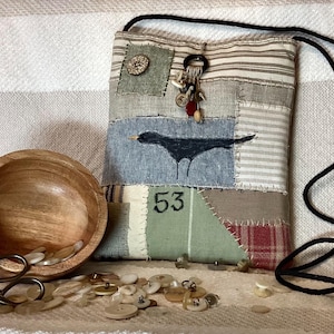 May include: A rectangular fabric crossbody bag featuring a patchwork design of striped and solid textiles in muted tones, a black bird illustration, the number 53, and decorative buttons. A wooden bowl filled with buttons and scissors are nearby.