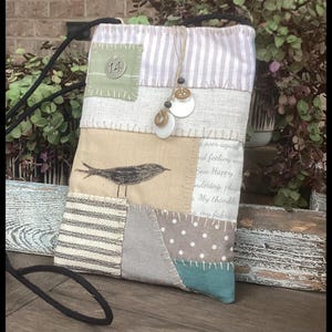 May include: A rectangular fabric crossbody bag featuring a patchwork design of beige, white, grey, and teal panels. The bag includes a black strap, a printed bird graphic, a green square patch with the number 14, and decorative white buttons.