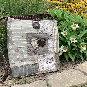 May include: A patchwork crossbody bag featuring a brown bird appliqué on a square fabric patch. The bag is constructed from various gray, white, and brown patterned fabrics, accented with multiple small white buttons and a large dark brown button closure.