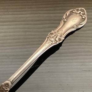 May include: A silver-toned metal spoon featuring an ornate handle with embossed floral detailing and a script letter R engraved near the top. The spoon has a smooth, oval-shaped bowl and a slender stem that connects to the decorative handle.