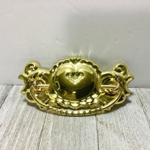May include: A shiny gold-colored drawer pull with a heart-shaped center and ornate detailing. The pull has a curved handle and decorative flourishes on either side, suitable for furniture restoration or crafting projects.