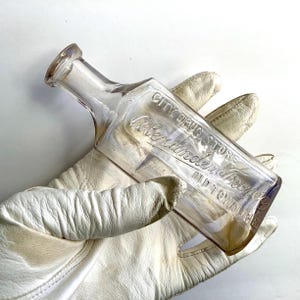 May include: A clear, antique glass bottle with a rectangular body and a narrow neck. The bottle has embossed text that reads "CITY DRUG STORE" and "Alexander & Frazee OLD TOWN ME". The bottle is held by a person wearing white leather gloves.