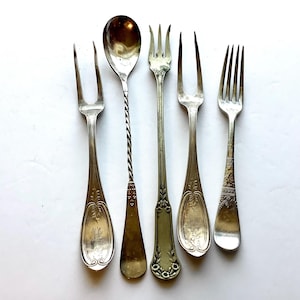 May include: An assortment of antique silverware, including forks and a spoon, arranged on a white surface. The silverware features ornate designs and engraved details. The spoon has a twisted handle. The forks have different tine configurations.