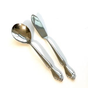 May include: A silver spoon and butter knife set. The spoon has a rounded bowl, and the butter knife has a flat, rounded blade. Both utensils have decorative handles with floral detailing. The set is made of a shiny, reflective metal.