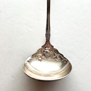 May include: Silver-colored serving ladle with a decorative grape and leaf design on the bowl. The handle is long and slender, with a detailed floral pattern near the bowl. The ladle has a curved bowl shape.