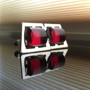 May include: Silver-toned cufflinks with a rectangular frame and a curved, rectangular, red-colored center. The cufflinks are highly reflective, with a mirrored surface. The design is simple and elegant.