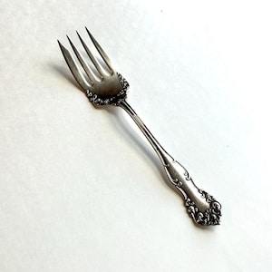 May include: A silver-toned metal fork featuring four tines and an ornate, embossed scrollwork pattern along the handle and the base of the tines. The metal surface displays a polished finish with intricate detailing throughout the entire piece.