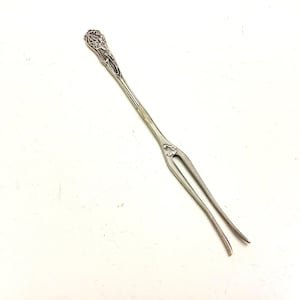 May include: A silver-toned, two-pronged fork with an ornate floral design on the handle and at the base of the tines. The fork is likely vintage or antique, and is set against a white background.