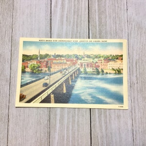 May include: Vintage postcard featuring the North Bridge over the Androscoggin River in Lewiston and Auburn, Maine. The image shows a bridge with cars, buildings, and a river. The postcard has a light cream border and the text "North Bridge over Androscoggin River, Lewiston and Auburn, Maine."