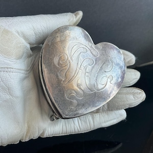 May include: A heart-shaped, silver-toned compact with an engraved floral design and initials. The compact is held in a white leather glove, showcasing its intricate details and vintage appeal. The compact is a vintage accessory.