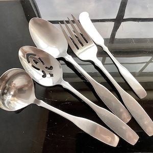 May include: A set of stainless steel silverware including a ladle, slotted spoon, serving spoon, fork, and butter knife. The utensils have a simple, elegant design with long, slender handles and are arranged on a reflective surface.