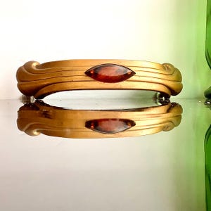 May include: A gold-toned bracelet with a central, oval-shaped, reddish-brown stone. The bracelet has a curved, Art Deco design with horizontal lines and a reflective surface. The bracelet is displayed on a reflective surface.