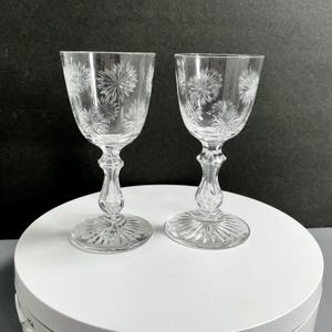 May include: Two clear, stemmed wine glasses with etched starburst designs. The glasses have a rounded bowl, a decorative stem, and a circular base. The glasses are displayed on a white surface against a dark background.