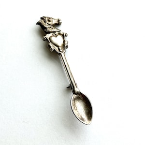 May include: A silver-toned spoon with a horse head design at the top. The spoon features a heart-shaped emblem and a small, oval bowl. The spoon is likely vintage and has a detailed, ornate design.