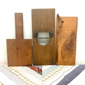 May include: Three vintage wooden kitchen tools including a paddle-shaped board, a rectangular slicer with a metal blade and serrated edge, and a rectangular board with a leather hanging loop. Each item features a natural brown wood grain finish.