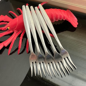 May include: A set of five silver-toned stainless steel seafood forks with slender, textured handles arranged in a fan shape. The forks are positioned in front of a bright red, segmented plastic lobster toy with visible black eyes.