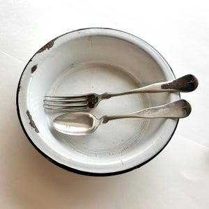 May include: A round white enamel bowl with a black rim and visible areas of chipped finish. Inside the bowl rests a silver-toned metal fork and a matching silver-toned metal spoon, both showing signs of wear and tarnishing on their surfaces.