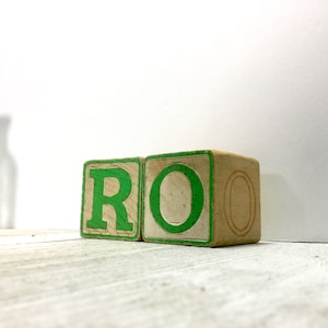 May include: Two wooden alphabet blocks with green painted letters spelling "RO". The blocks are beige with green trim and the letters are in a bold, sans-serif font. The blocks are on a white surface.