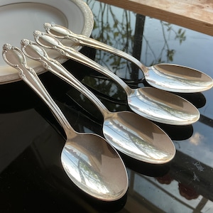 May include: A set of four polished silver-tone metal spoons arranged diagonally. Each spoon features a slender handle with a decorative embossed scroll pattern at the top and a smooth, oval-shaped bowl. The metal surface is highly reflective.