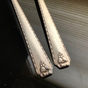May include: Two antique silver-colored spoons with a decorative grape and leaf design at the base of the handle. The spoons have a detailed border pattern and are set against a dark background.