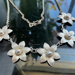 May include: Silver-toned necklace featuring six detailed flower pendants. Each flower has a textured design and a central, raised element. The necklace chain is delicate, with a hook clasp. The jewelry is displayed on a dark, reflective surface.