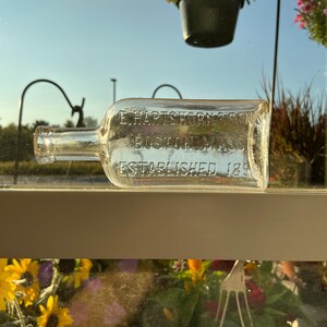 May include: A clear, rectangular glass bottle with a long neck, lying horizontally. The bottle has embossed text: "E. HARTSHORN & SONS, BOSTON, MASS., ESTABLISHED 1850." The bottle is positioned on a window sill, with a blurred background of flowers and greenery.