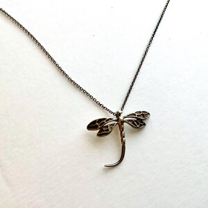 May include: A silver-toned dragonfly pendant necklace. The dragonfly has detailed wings and a curved body. The chain is a delicate, dark silver color. The necklace is displayed on a white background.
