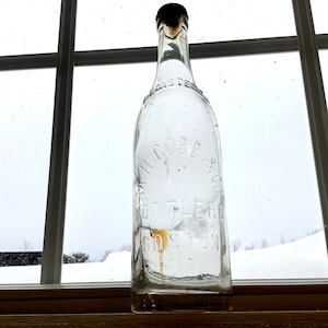 May include: Clear glass bottle with a dark cap, featuring embossed text that reads "H. COBE & CO. BOTTLERS BOSTON". The bottle is positioned in front of a window with a snowy outdoor scene visible.