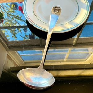 May include: A silver-toned metal soup ladle featuring a long, slender handle with a decorative shell-like motif at the top. The ladle has a deep, rounded bowl designed for serving liquids, with a polished finish that reflects light across its surface.